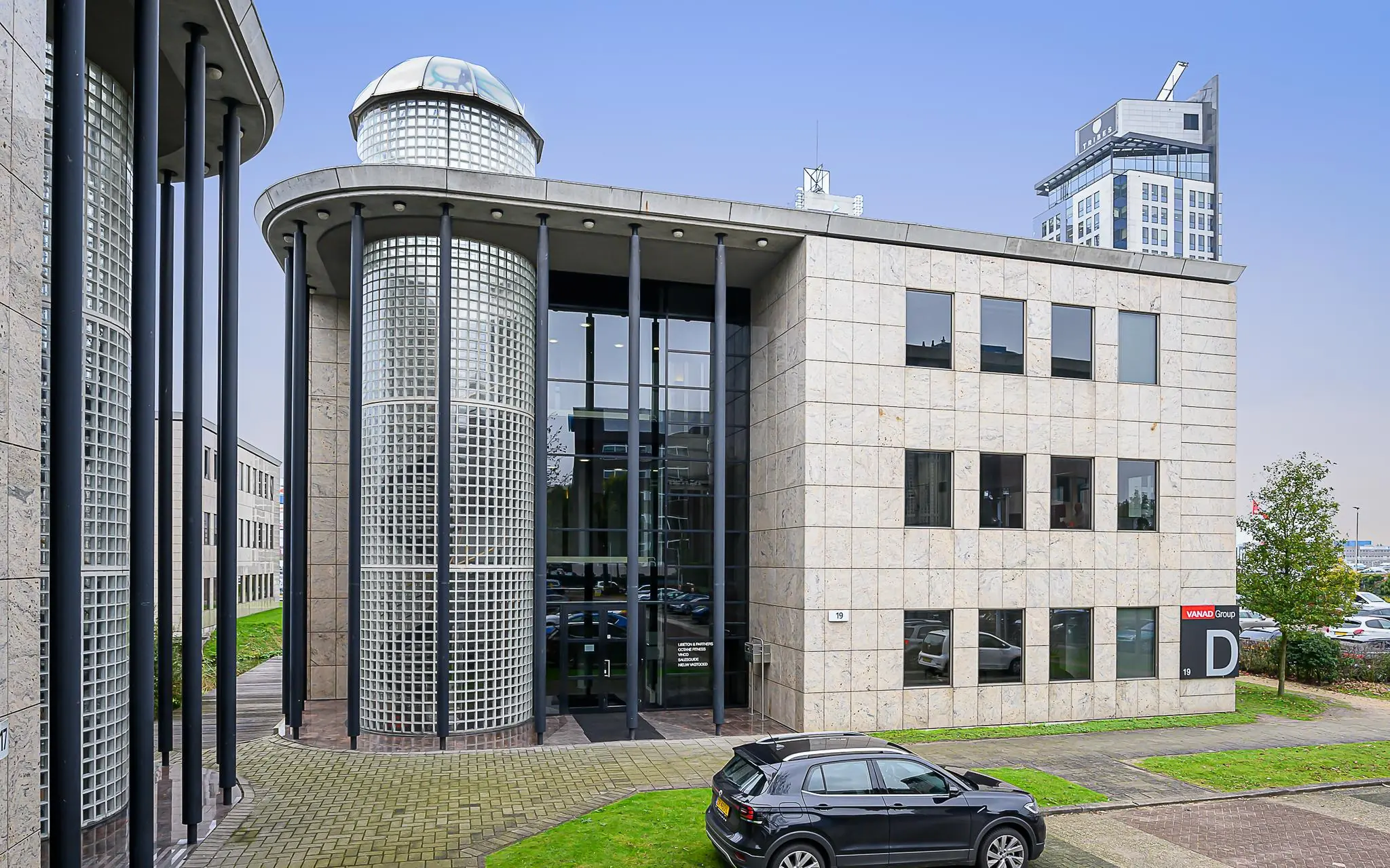 Modern kantoorgebouw aan de Rivium Westlaan 19 met glazen gevelpartijen en pilaren, met een auto geparkeerd voor de ingang.